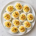 A platter of deviled eggs with pale yellow filling, paprika and chives